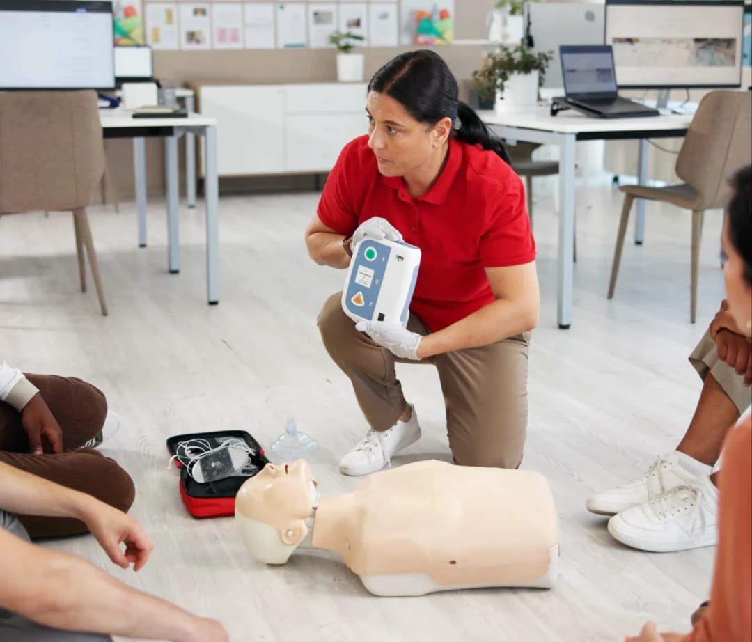 A first aid instructor in a red uniform demonstrating the use of an emergency medical device during an initial onboarding and skills assessment session.