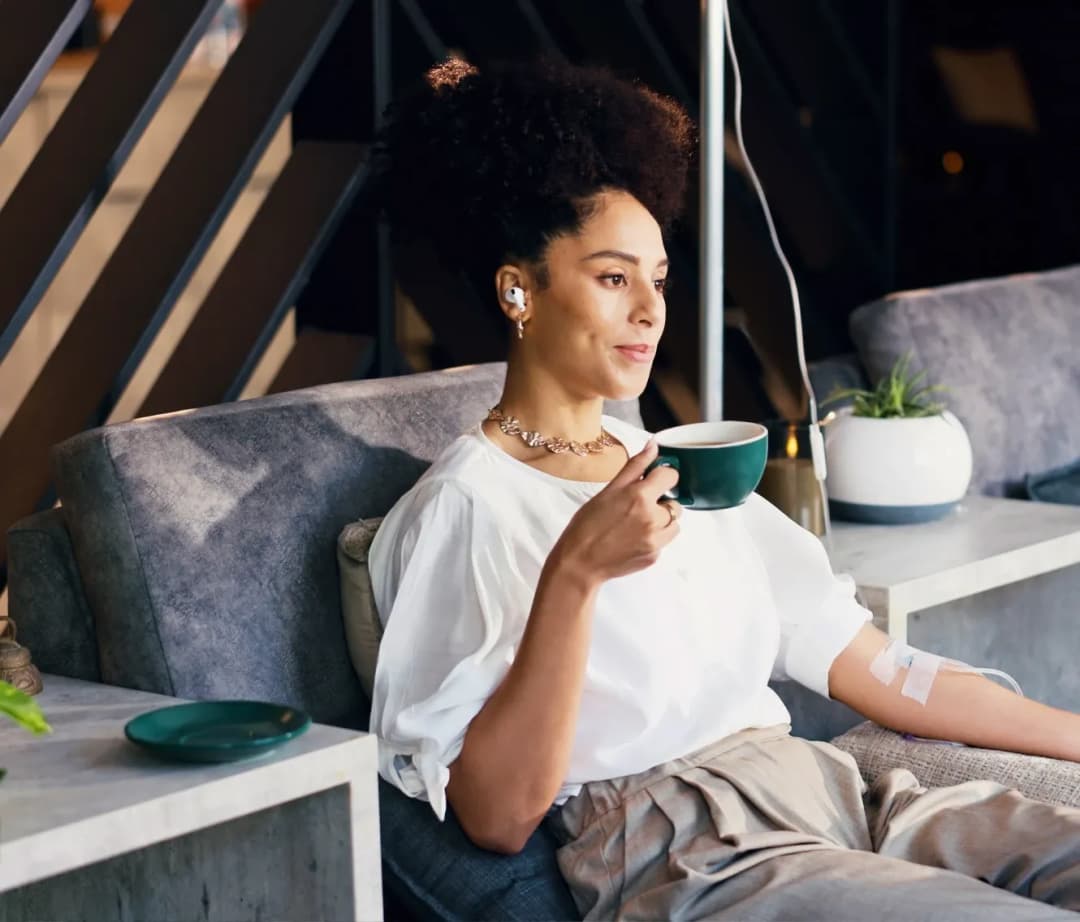 A relaxed woman smiling and enjoying a hot beverage on a sofa, illustrating the comfort and convenience of receiving on-demand home wellness care within 30 minutes.
