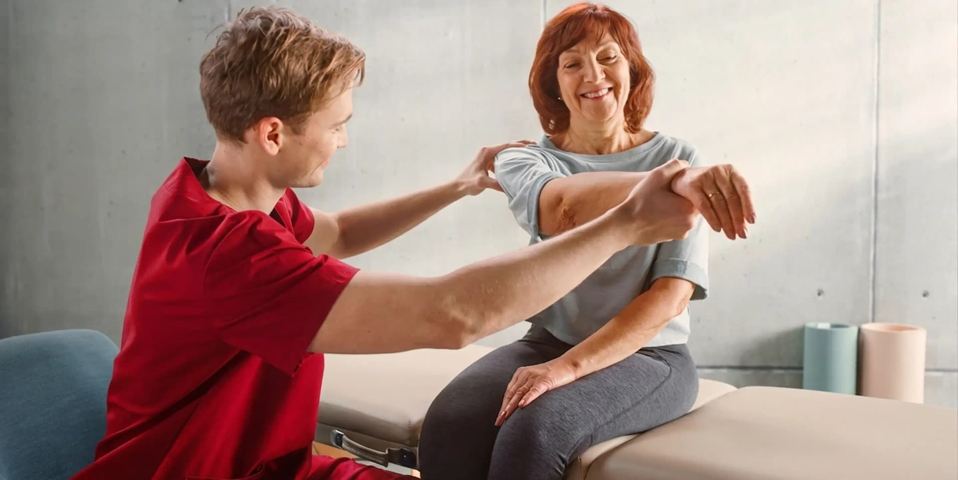 A male physiotherapist assisting a smiling elderly woman with upper-body exercises during a home musculoskeletal physiotherapy session by First Response Healthcare in Dubai.