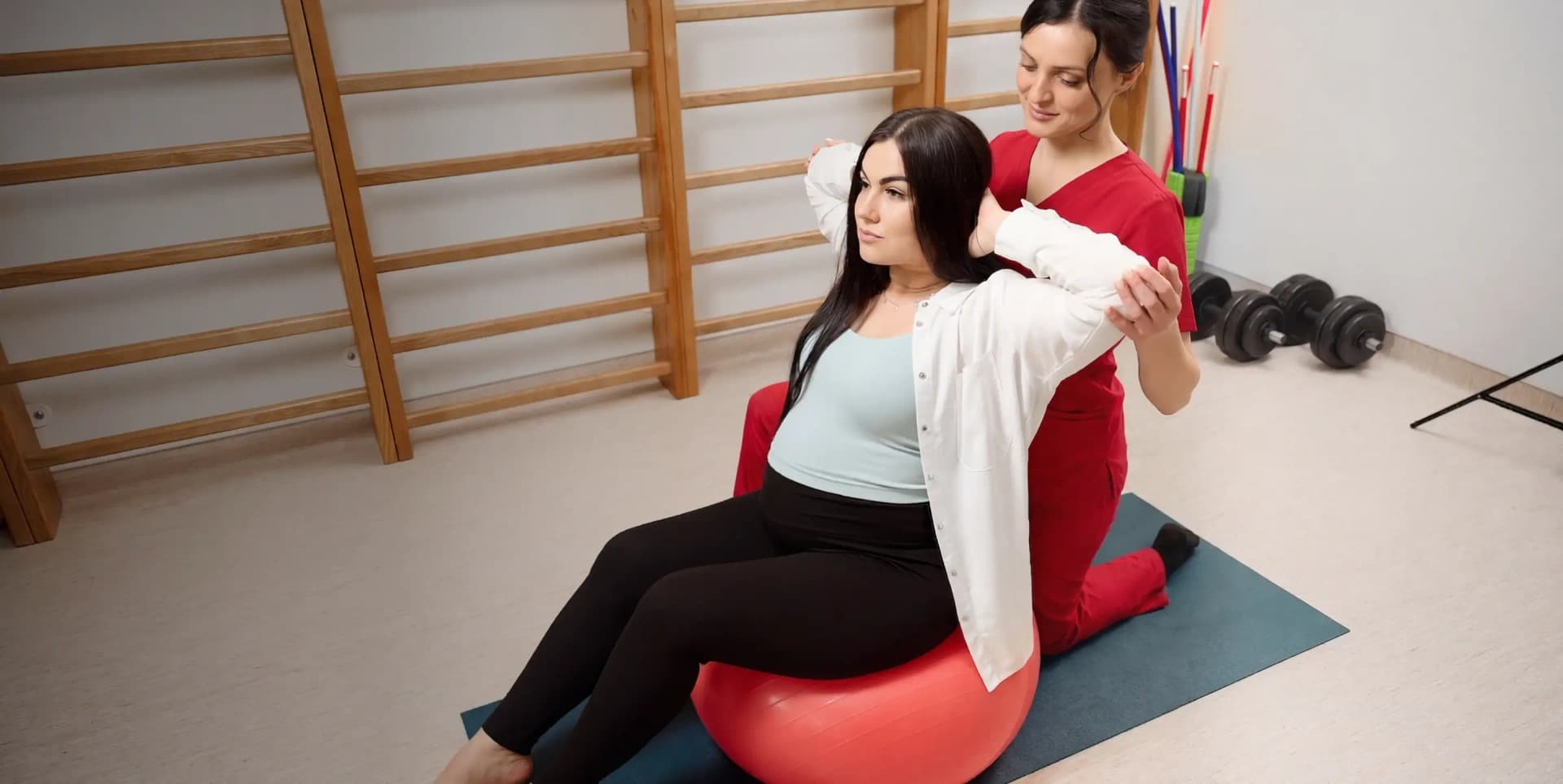 A female physiotherapist in uniform helping a woman with prenatal stability exercises on a fitness ball during a home session in Dubai.