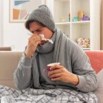 A man sitting on the couch indicating he has a flowing nose due to a cold