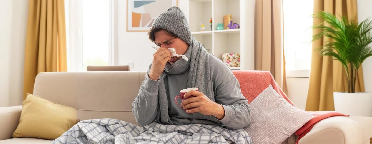 A man sitting on the couch indicating he has a flowing nose due to a cold