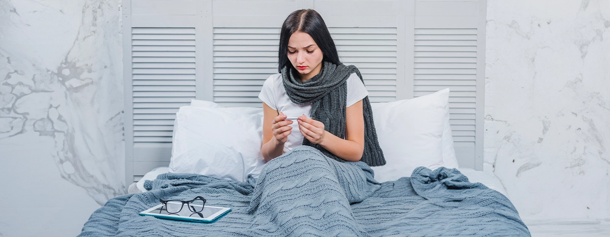 A woman on bed checking body temperature with a thermometer