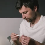 A man sitting on bed checking his temperature with a thermometer