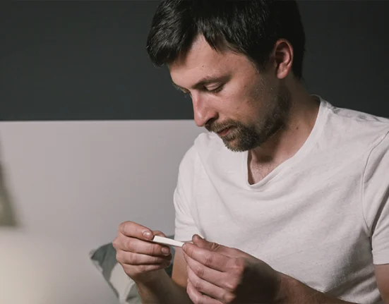 A man sitting on bed checking his temperature with a thermometer