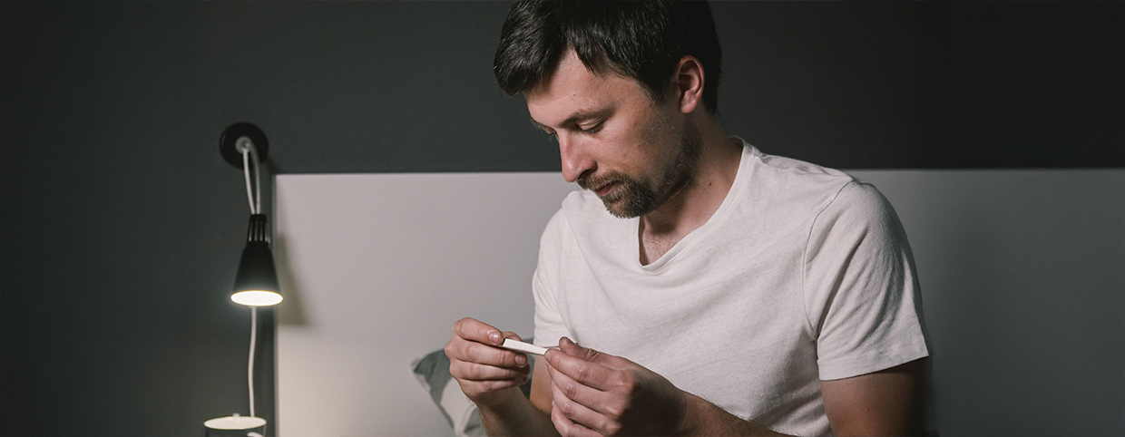 A man sitting on bed checking his temperature with a thermometer
