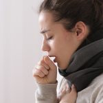 A woman coughing while sitting on a bed with a scarf around her neck