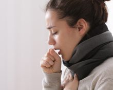 A woman coughing while sitting on a bed with a scarf around her neck