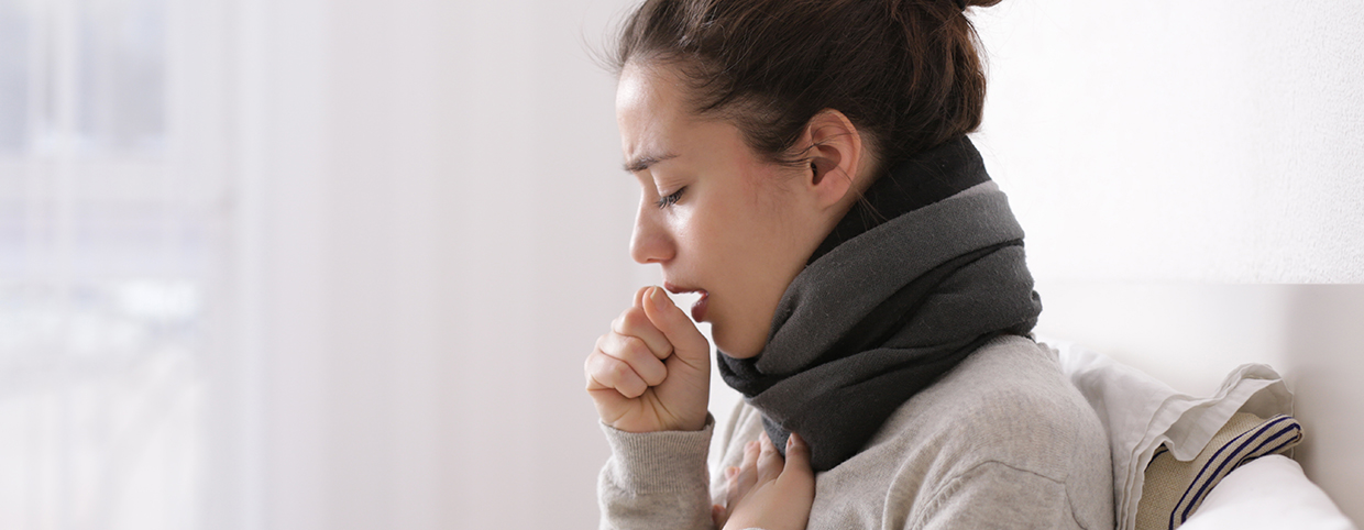 A woman coughing while sitting on a bed with a scarf around her neck