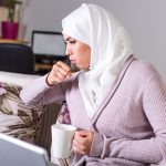 A woman coughing while sitting on a couch with a laptop and holding a mug