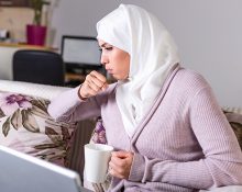 A woman coughing while sitting on a couch with a laptop and holding a mug