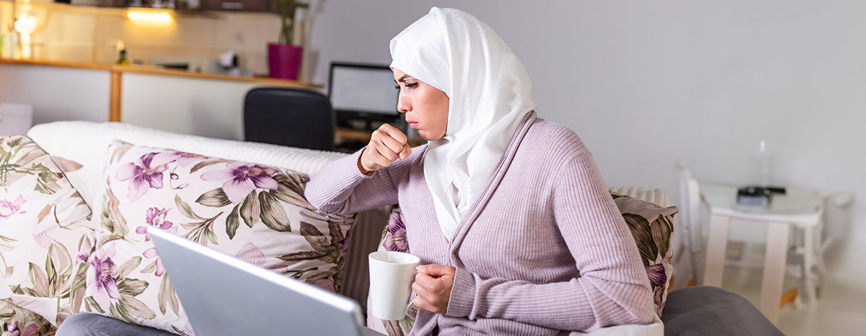 A woman coughing while sitting on a couch with a laptop and holding a mug